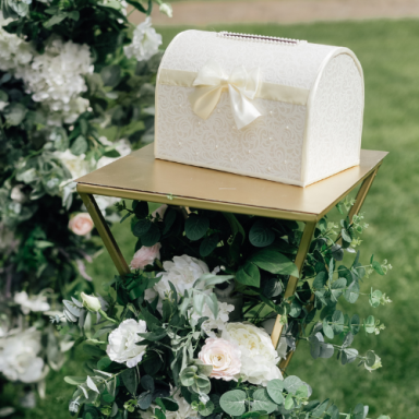Boîte blanche ornée sur un support doré, entourée de fleurs blanches et vertes.