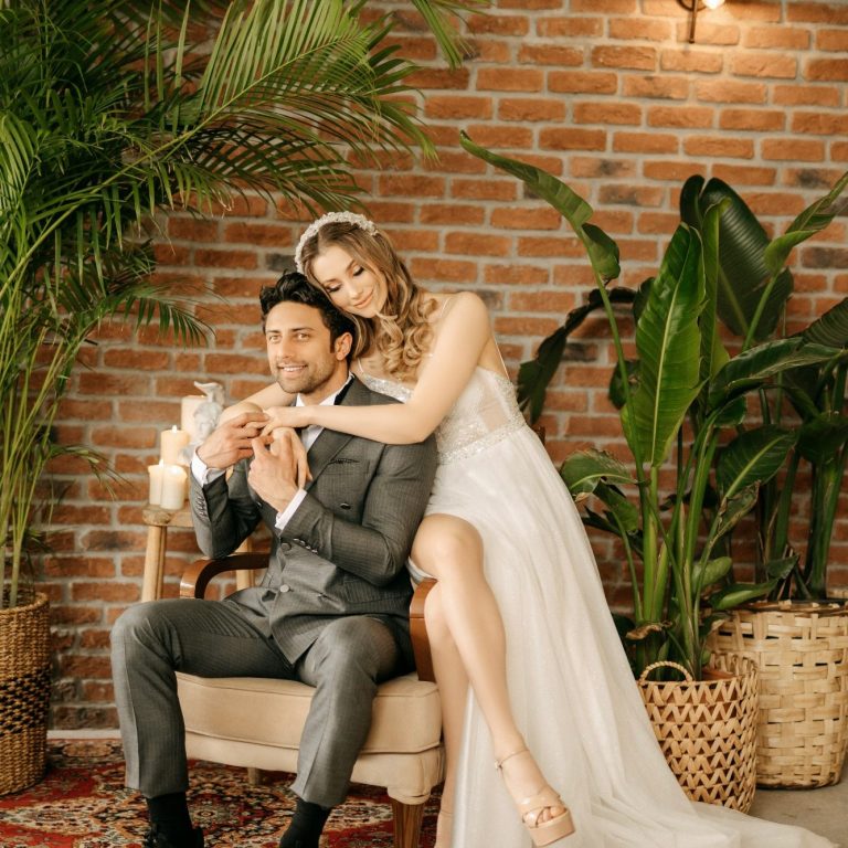 Un couple heureux en tenue de mariage, assis dans un décor de briques et de plantes.