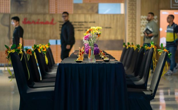Table dressée avec des fleurs colorées, chaises noires, et personnes en arrière-plan.