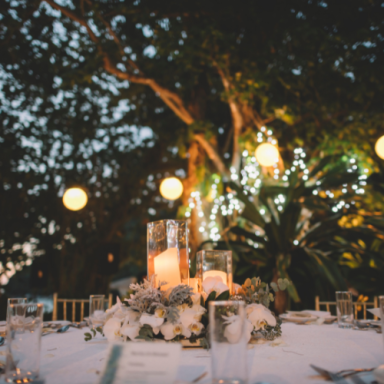 Table set for an outdoor event, adorned with candles and greenery under soft lighting.