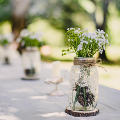 Des pots en verre avec des fleurs blanches, décorés de jute, sur une table en extérieur.