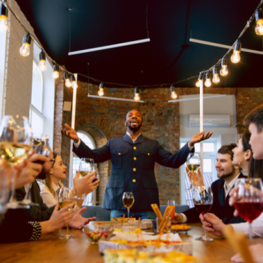 Homme souriant accueille des invités autour d'une table garnie de plats et boissons.